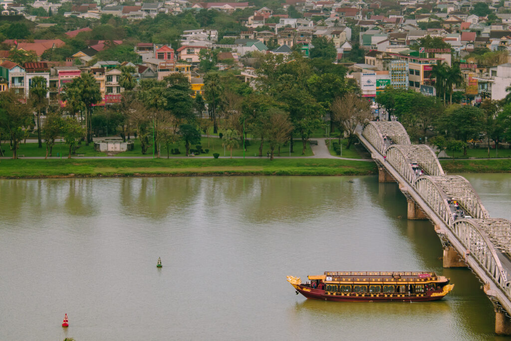 Ngự Thuyền Hueritage trên sông Hương cạnh cầu Trường Tiền, phía sau là hàng cây ven bờ và phố Huế.